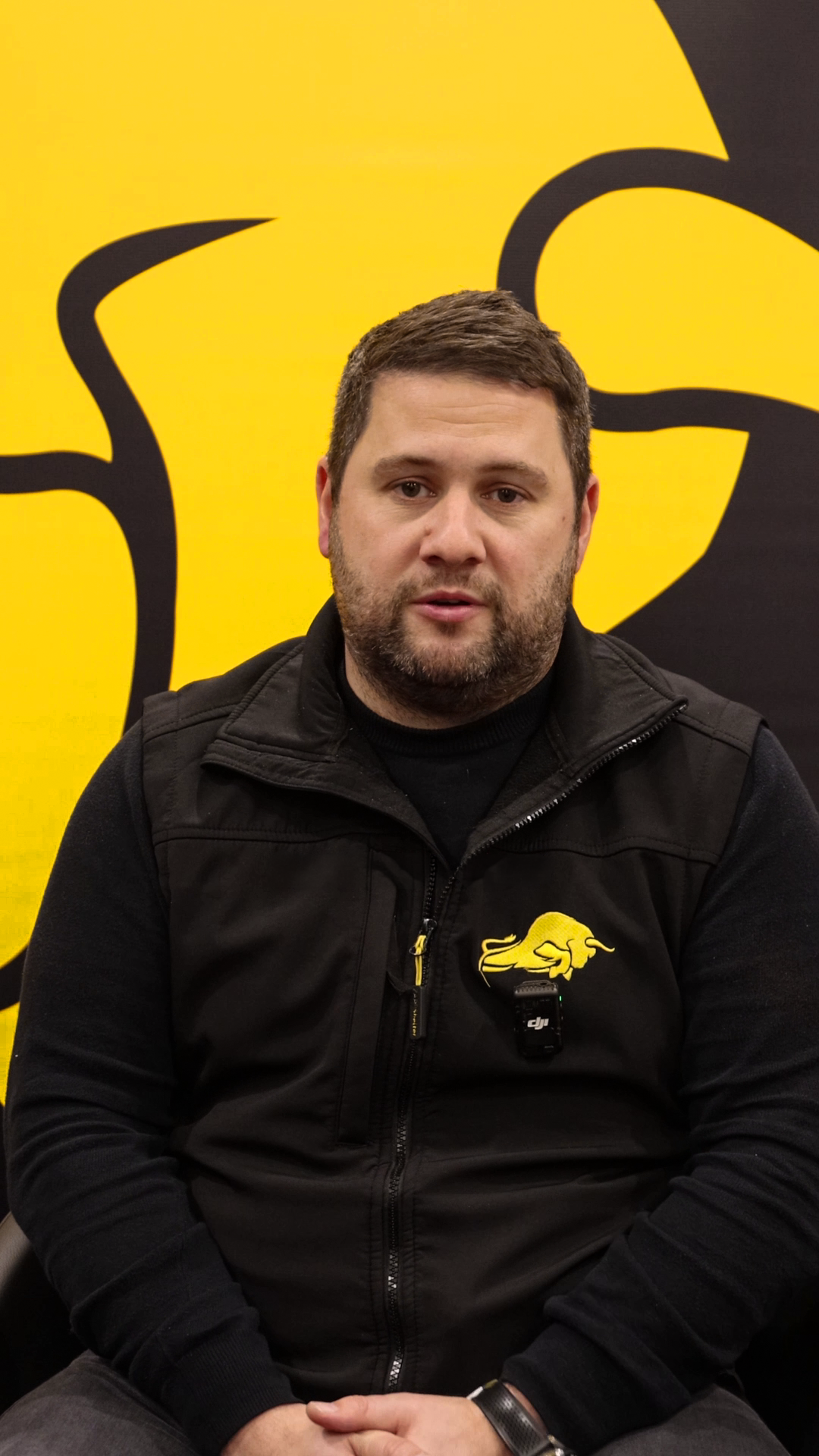 Video still: Liam speaking in front of a yellow and black branded background.