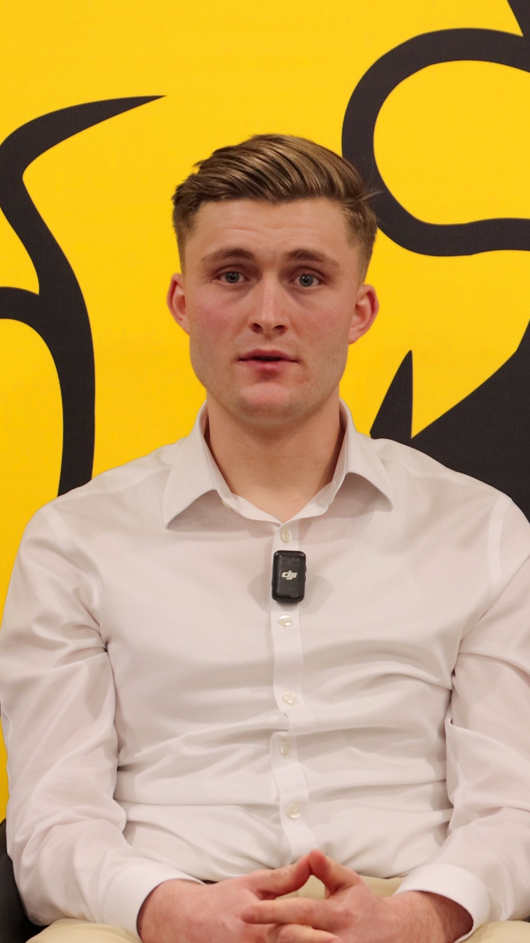 Video still: Ash speaking in front of a yellow and black branded background.