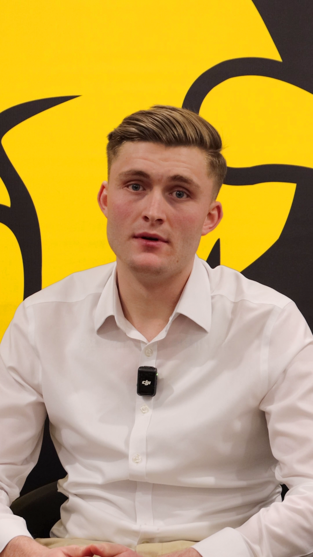 Video still: Ash speaking in front of a yellow and black branded background.
